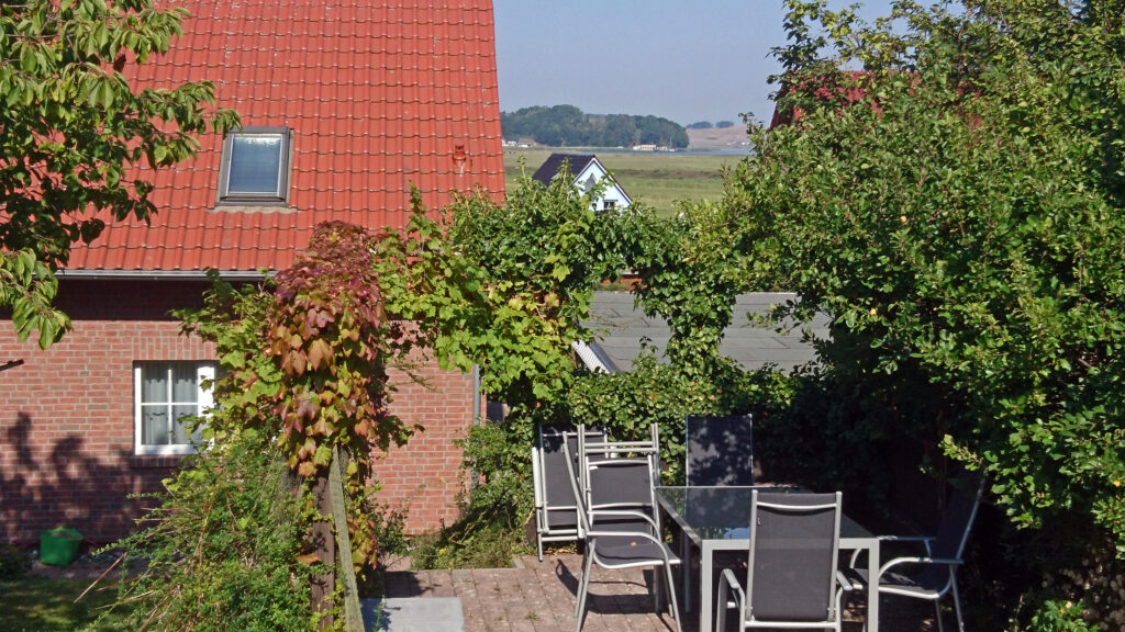 Ein gemütlicher Garten mit einem Tisch und sechs Stühlen aus Metall. Der Tisch hat eine Glasplatte. Im Hintergrund ist ein rotziegeliges Haus mit einem Dachfenster zu sehen. Rechts vom Haus stehen hohe Büsche und Bäume, die einen Teil des Gartens abschirmen. Im Hintergrund ist ein See mit Booten zu erkennen.